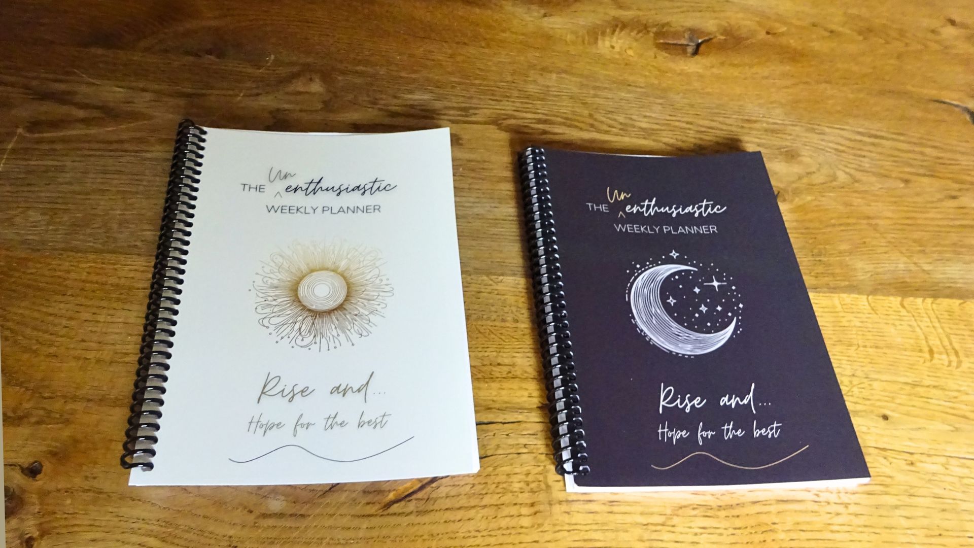 Photograph of two copies of The Unenthusiastic Weekly Planner "Rise and hope for the best", light sun and dark moon style covers, coil bound, laying side by side on a table.