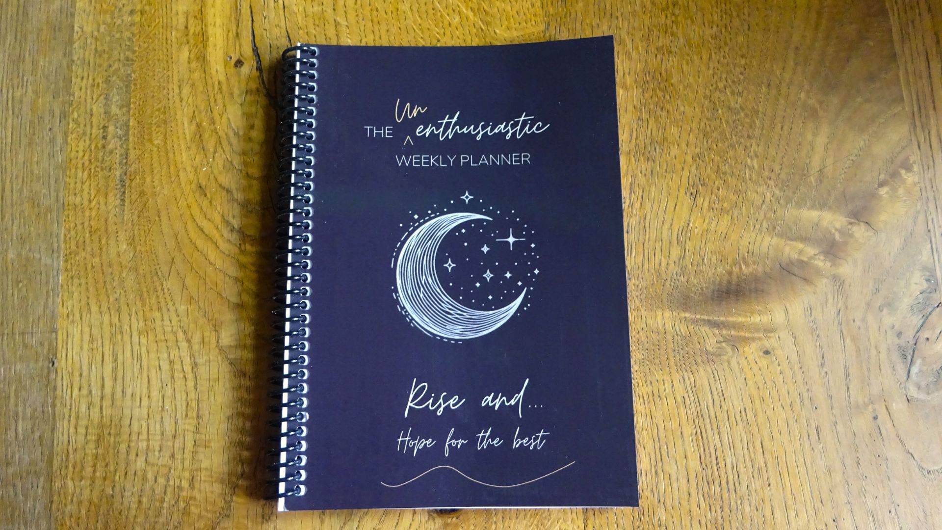 Photograph of The Unenthusiastic Weekly Planner "Rise and hope for the best", dark moon style cover, coil bound, laying on a table.