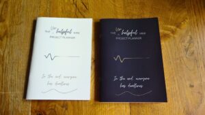 Photo of two copies The Unhelpful Mini Project Planner. light and dark cover editions, Saddle Stitched book laying on a table