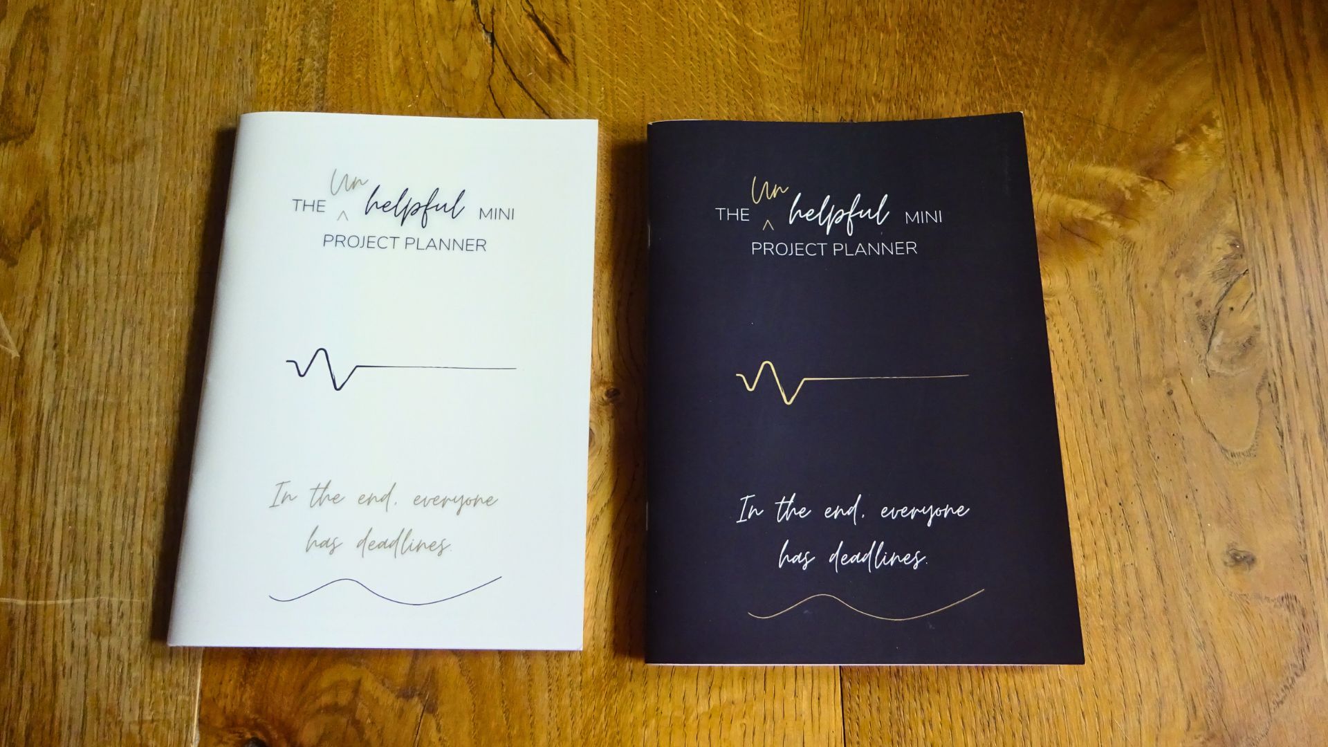 Photo of two copies The Unhelpful Mini Project Planner. light and dark cover editions, Saddle Stitched book laying on a table