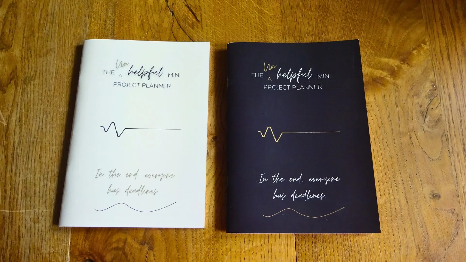 Photo of two copies The Unhelpful Mini Project Planner. light and dark cover editions, Saddle Stitched book laying on a table