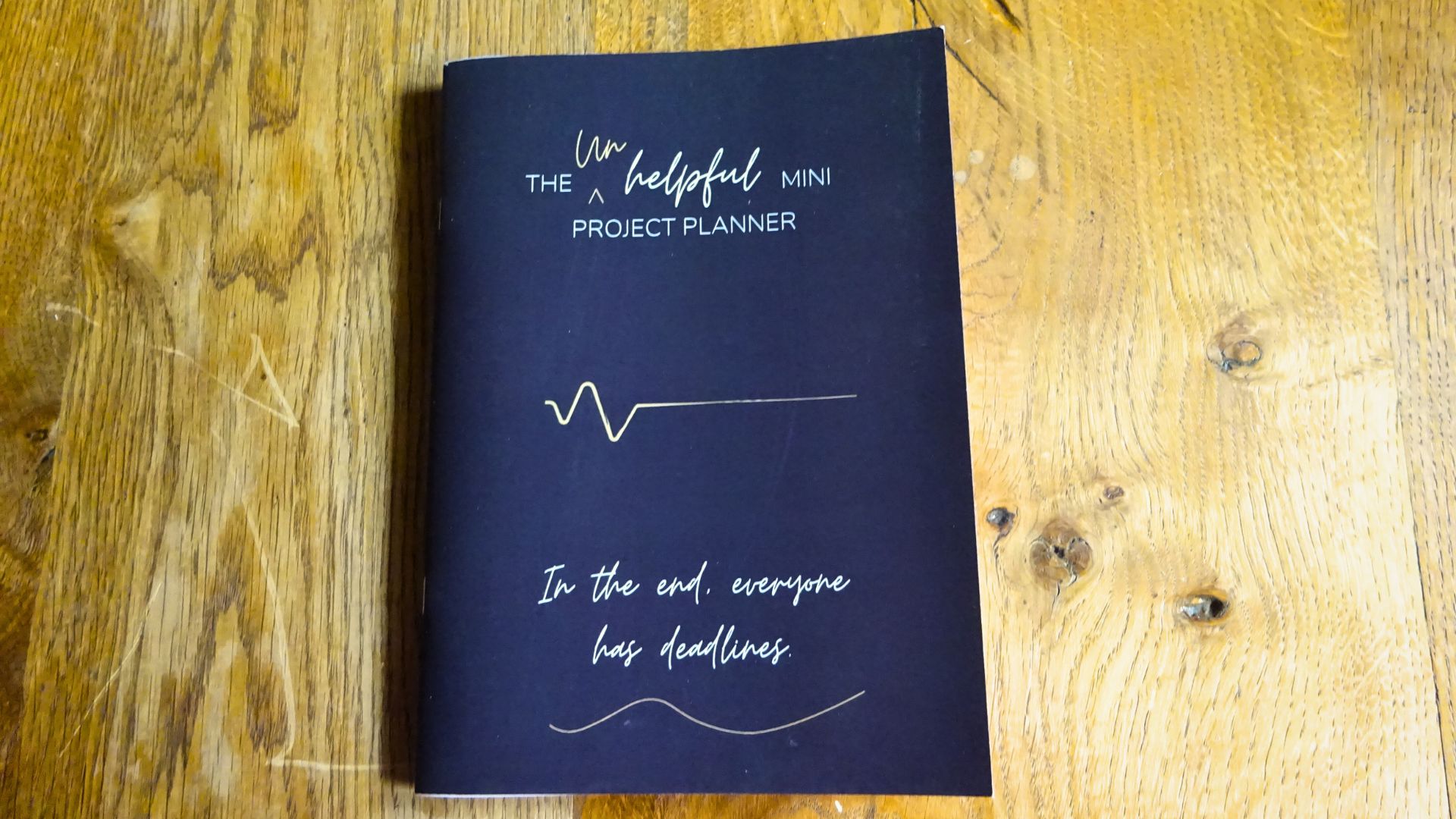 Photo of The Unhelpful Mini Project Planner Saddle Stitched book dark cover laying on a table
